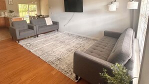 Uma sala de estar espaçosa com um amplo sofá cinzento, uma poltrona cinzenta e uma mesa de centro de madeira. Uma televisão de tela plana está fixada na parede, e há um ventilador de teto.