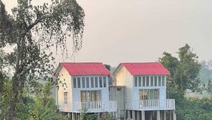 A two-story house with a red roof, elevated on stilts, surrounded by dense greenery.