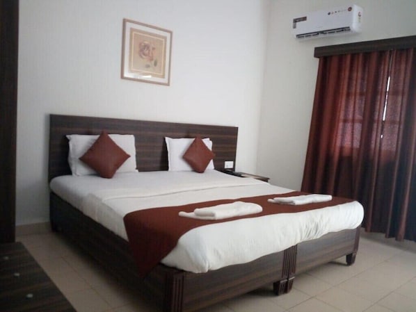 A hotel room with a large bed, a headboard, a wall-mounted air conditioner, and a window with curtains.
