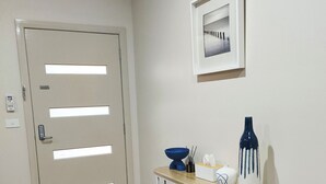 Un corridoio con pavimento in legno, una porta bianca con tastierino numerico, una piccola mensola bianca con un vaso e un libro, e un quadro appeso al muro.