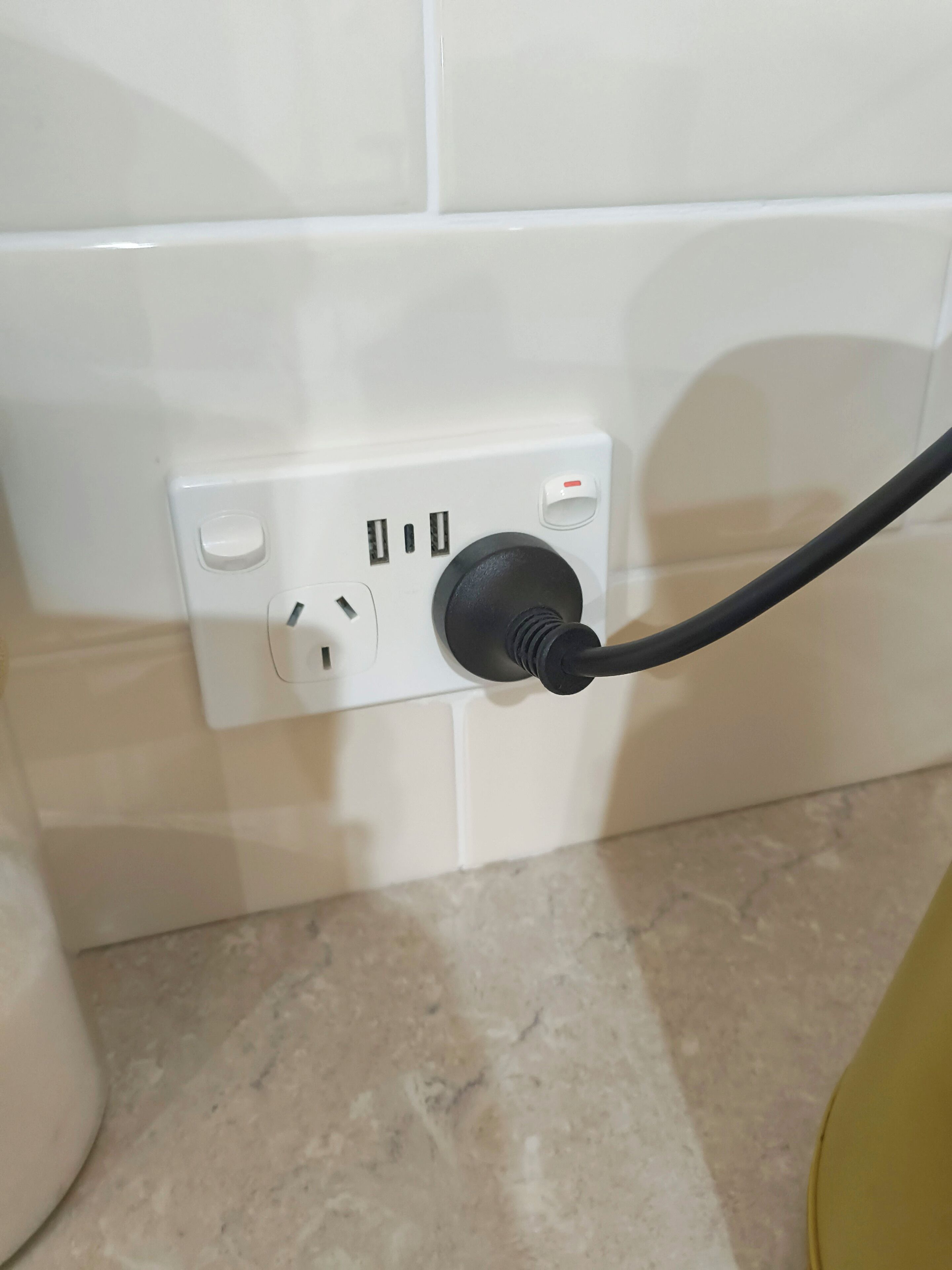 Una presa elèctrica blanca amb un endoll negre, davant d'una paret revestida de rajoles.