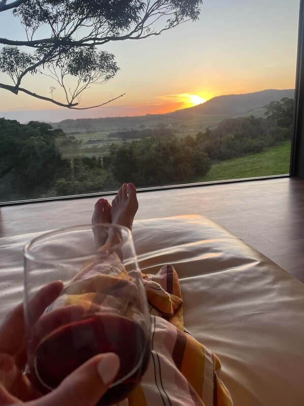 Une chambre avec un lit, un verre de vin rouge et une vue sur un paysage baigné par les couleurs d’un coucher de soleil.