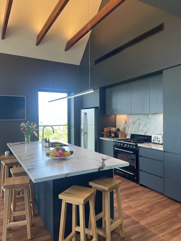 Een moderne keuken met een groot eiland, houten krukken, een inbouwoven en een roestvrijstalen koelkast.