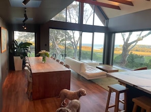 Un intérieur moderne avec une table à manger en bois, un canapé blanc et de grandes fenêtres donnant sur l’extérieur.