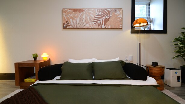 A neatly made bed with a green bedspread, a bedside lamp, a small plant on the nightstand, and a large abstract painting on the wall. - Bluff Street Escape • Modern 1BR Near Downtown (Dubuque)
