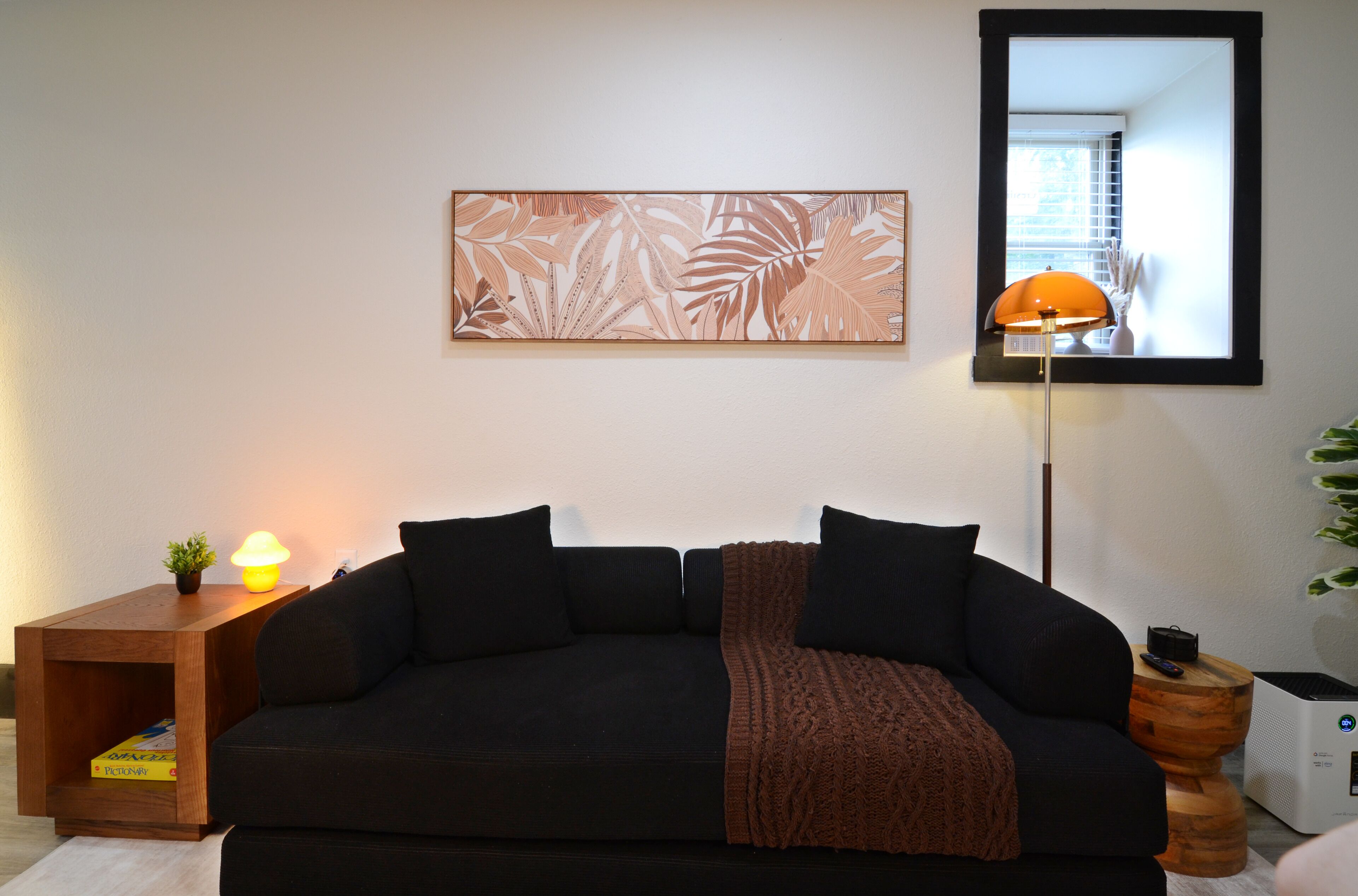 En stue med en mørk sofa, et lille bord med en lampe, et billede i ramme på væggen og et stort spejl der viser et vindue.