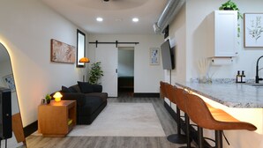 A modern living room with a sofa, a coffee table, and a TV. There is a kitchen area with a counter and stools. - Bluff Street Escape • Modern 1BR Near Downtown (Dubuque)