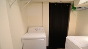 A compact laundry room with a front-loading washer and dryer, a black shower curtain, and a shelf with cleaning supplies. - Bluff Street Escape • Modern 1BR Near Downtown (Dubuque)