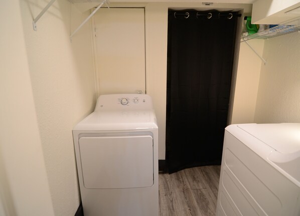 Une petite buanderie équipée d’un lave-linge et d’un sèche-linge à chargement frontal, d’un rideau de douche noir et d’une étagère contenant des produits de nettoyage.