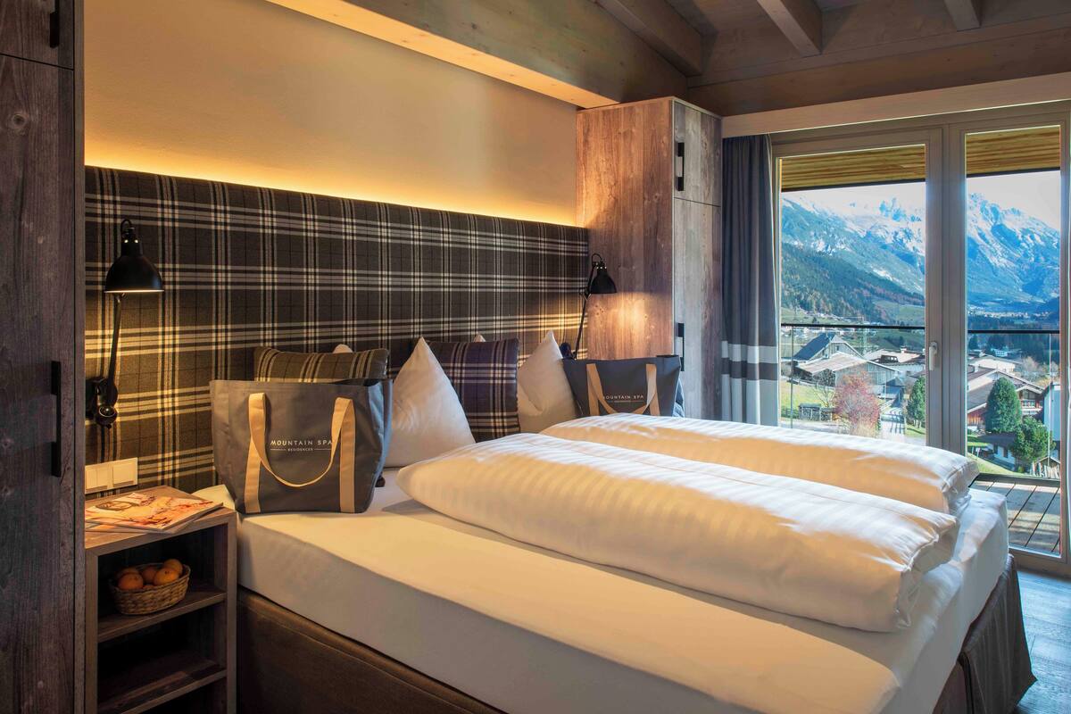 Habitación de hotel con cama, cabecera de cuadros, mesita de noche con lámpara, cesta de naranjas y vista a las montañas por la ventana.
