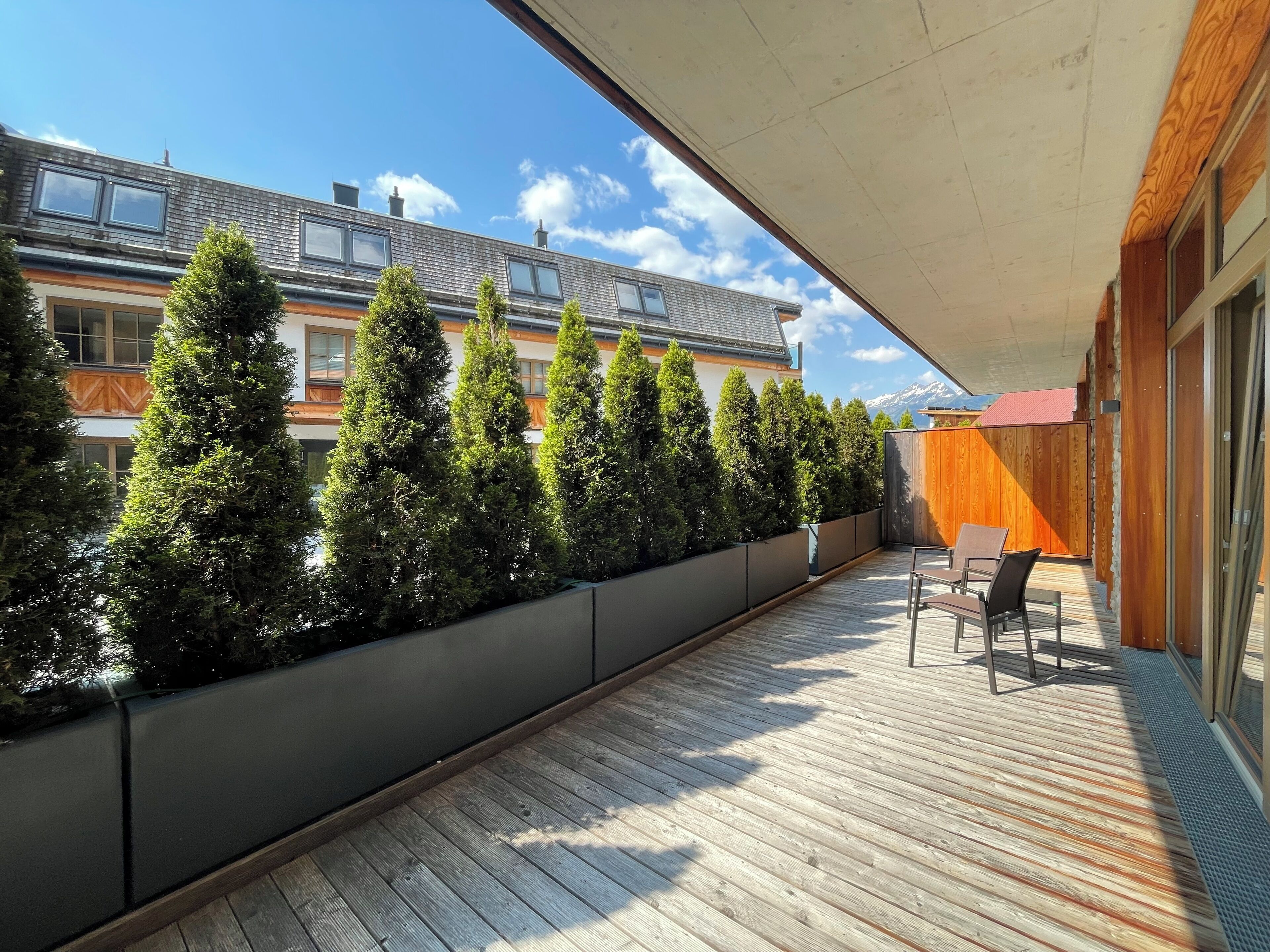 Una terraza de madera con un hilera de árboles cuidadosamente podados, una zona de estar con sillas y vistas a un edificio de varios pisos con balcones.