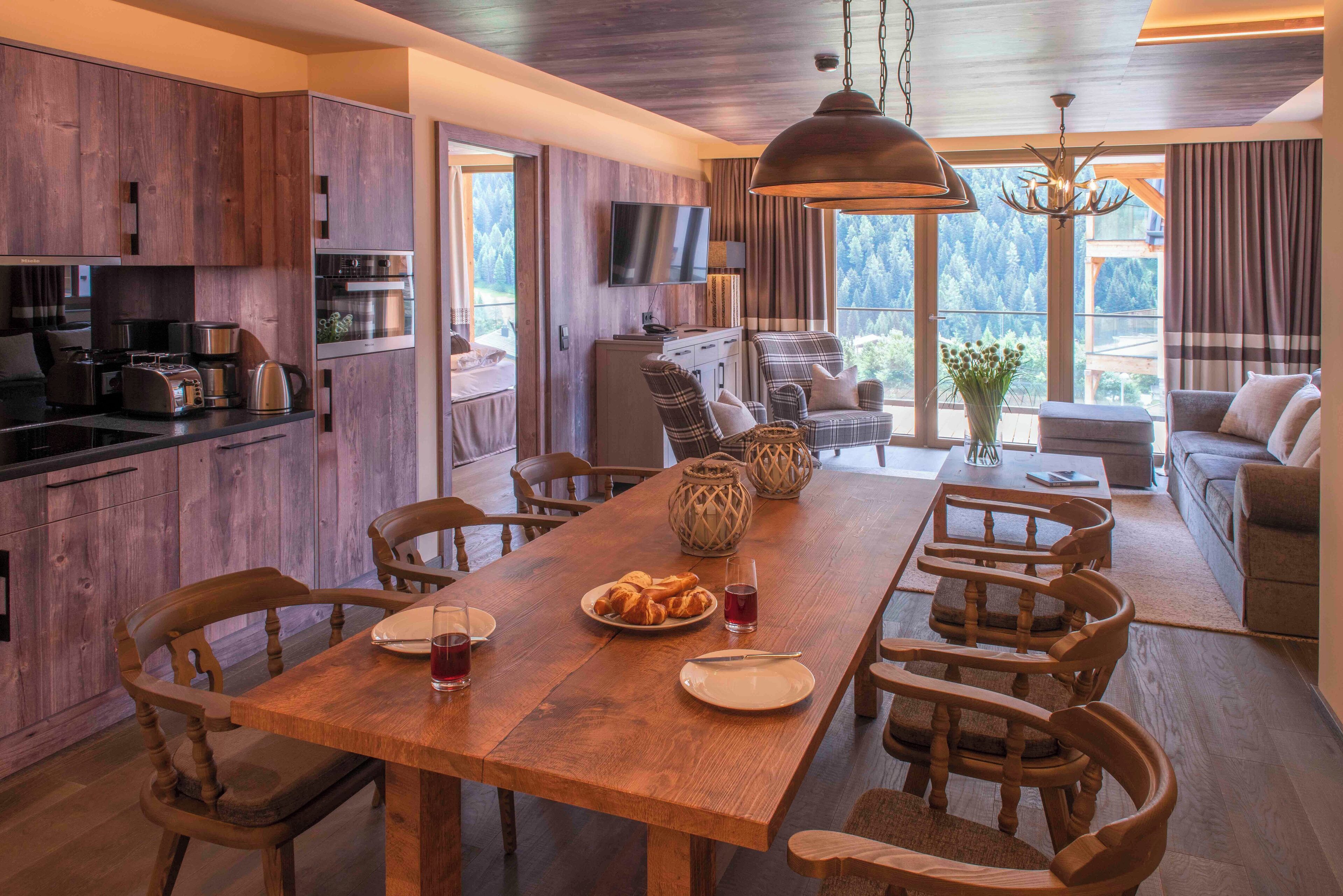 A modern kitchen with wooden cabinetry, a dining area with a wooden table and chairs, a television mounted on the wall, and a view of the outdoors through large windows.