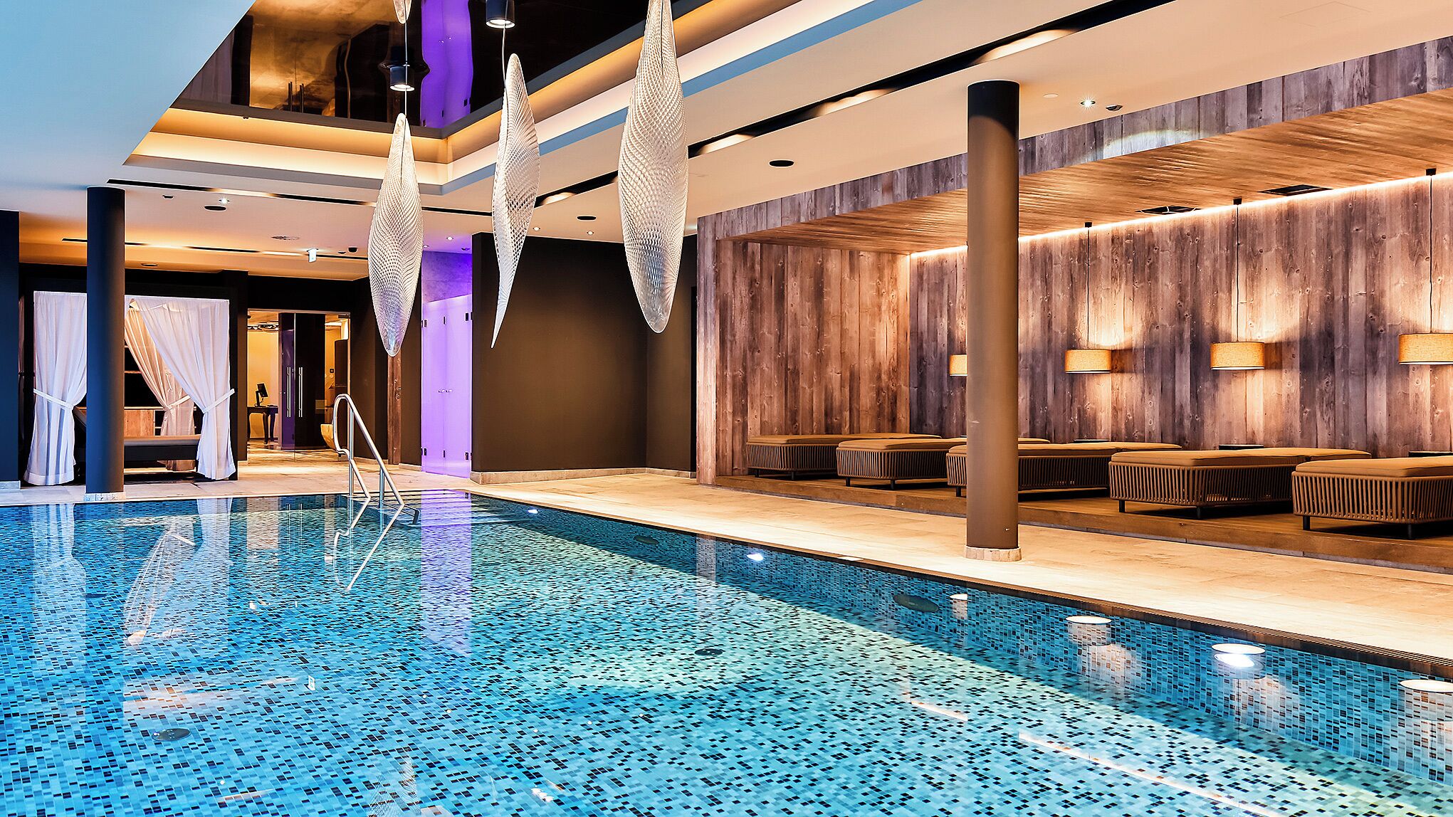 A modern indoor pool area with a clear pool, seating along the sides, and a wooden wall backdrop.