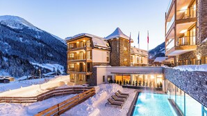 Zona de piscina exterior cubierta de nieve con sillas de descanso, una terraza de madera y un edificio con múltiples balcones y ventanas grandes.