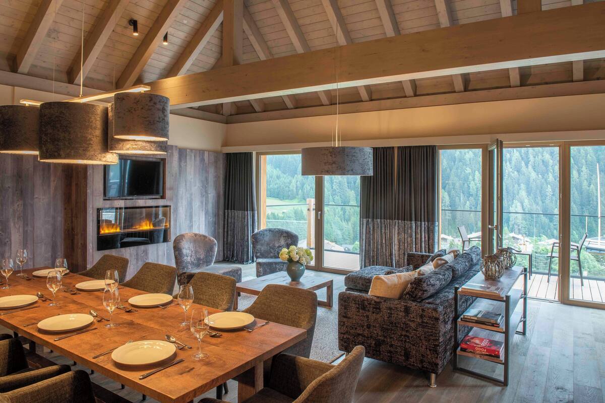 Un comedor con una mesa de madera dispuesta para una comida, rodeado de asientos cómodos y ventanas grandes con vistas al exterior.