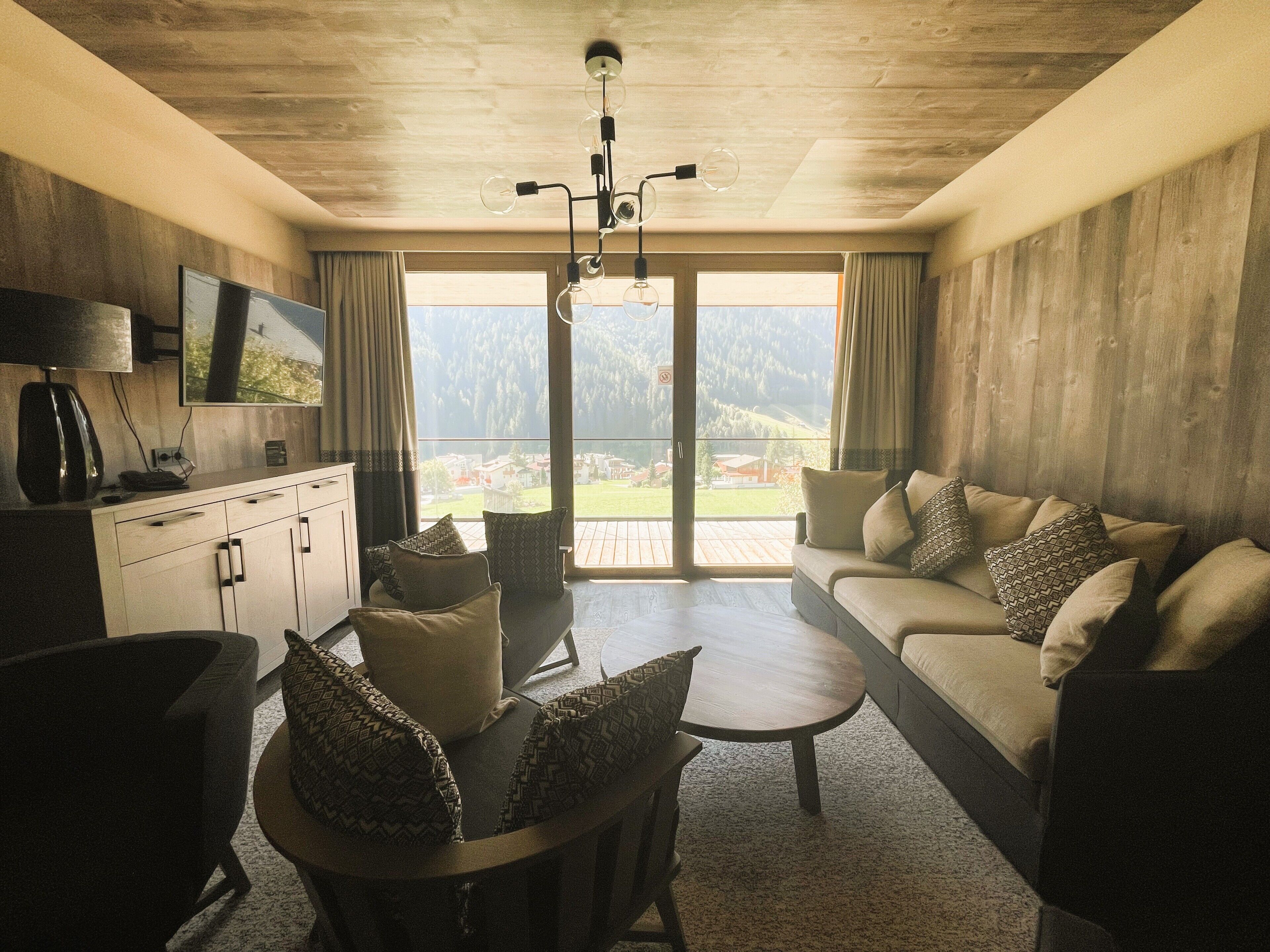A modern living room with a sofa, coffee table, and chairs, a TV on the wall, and a view of the outdoors.