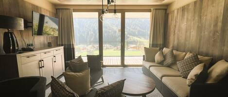 A modern living room with a sofa, coffee table, and chairs, a TV on the wall, and a view of the outdoors.