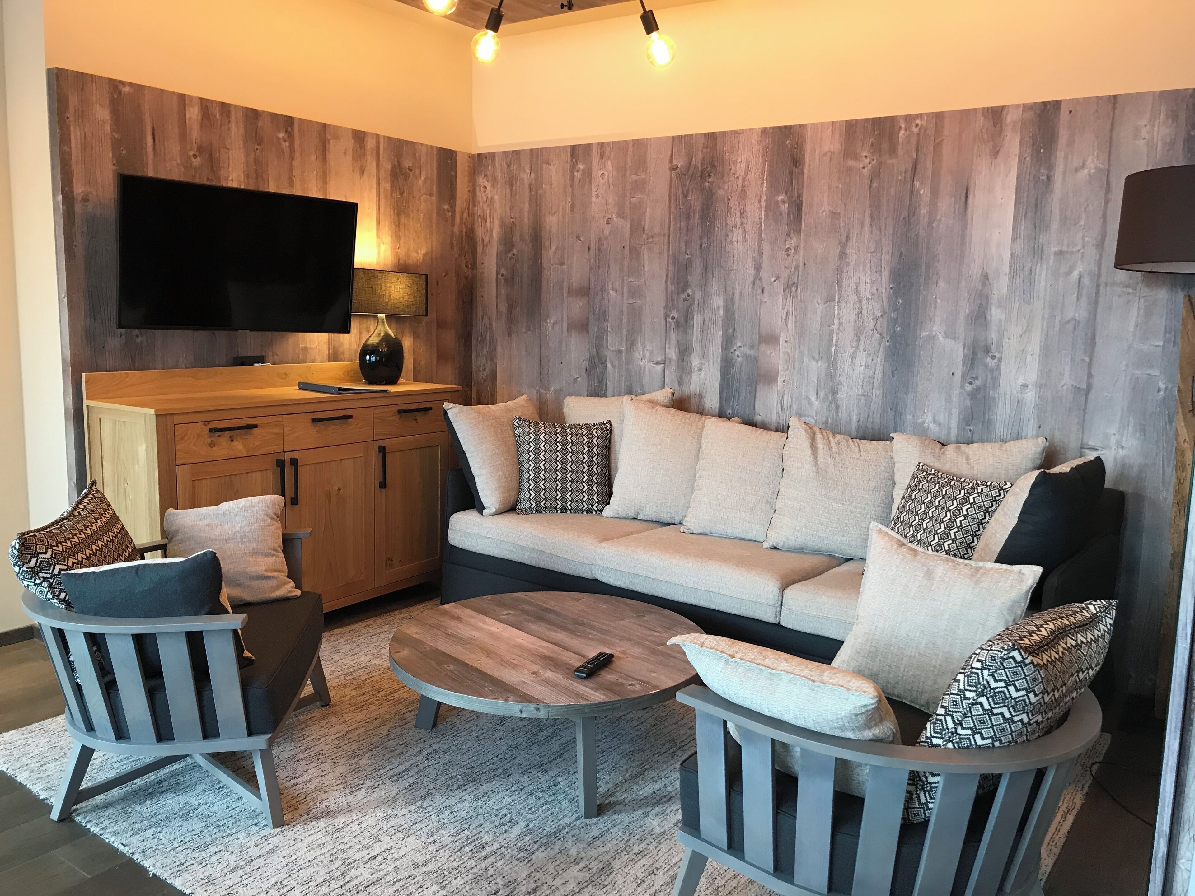 Een gezellige zithoek met een houten accentmuur, een flatscreen televisie, een bank met opvallende kussens, een salontafel en een fauteuil.