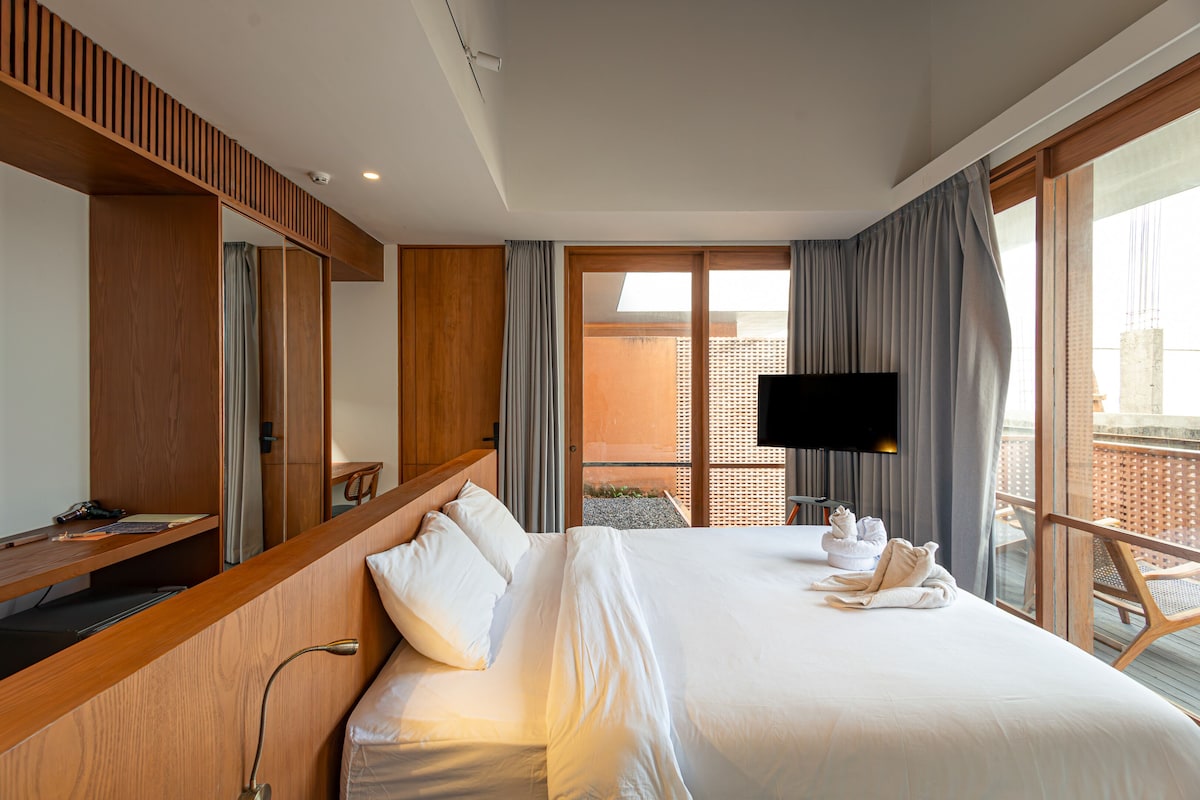 A hotel room with a large bed, a desk, a flat-screen TV, and a balcony with a view of the city.
