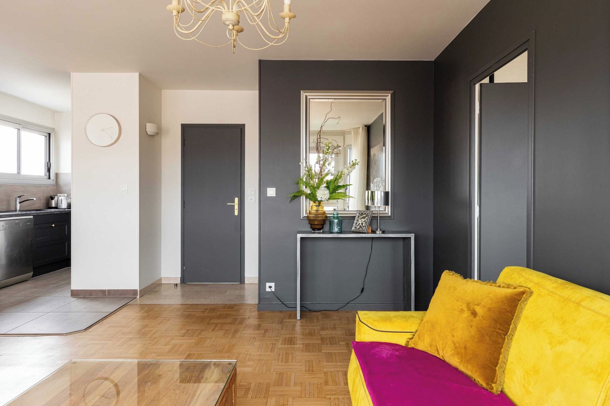 Een moderne woonkamer met een houten vloer, een gele bank, een console met een vaas en een grote spiegel.