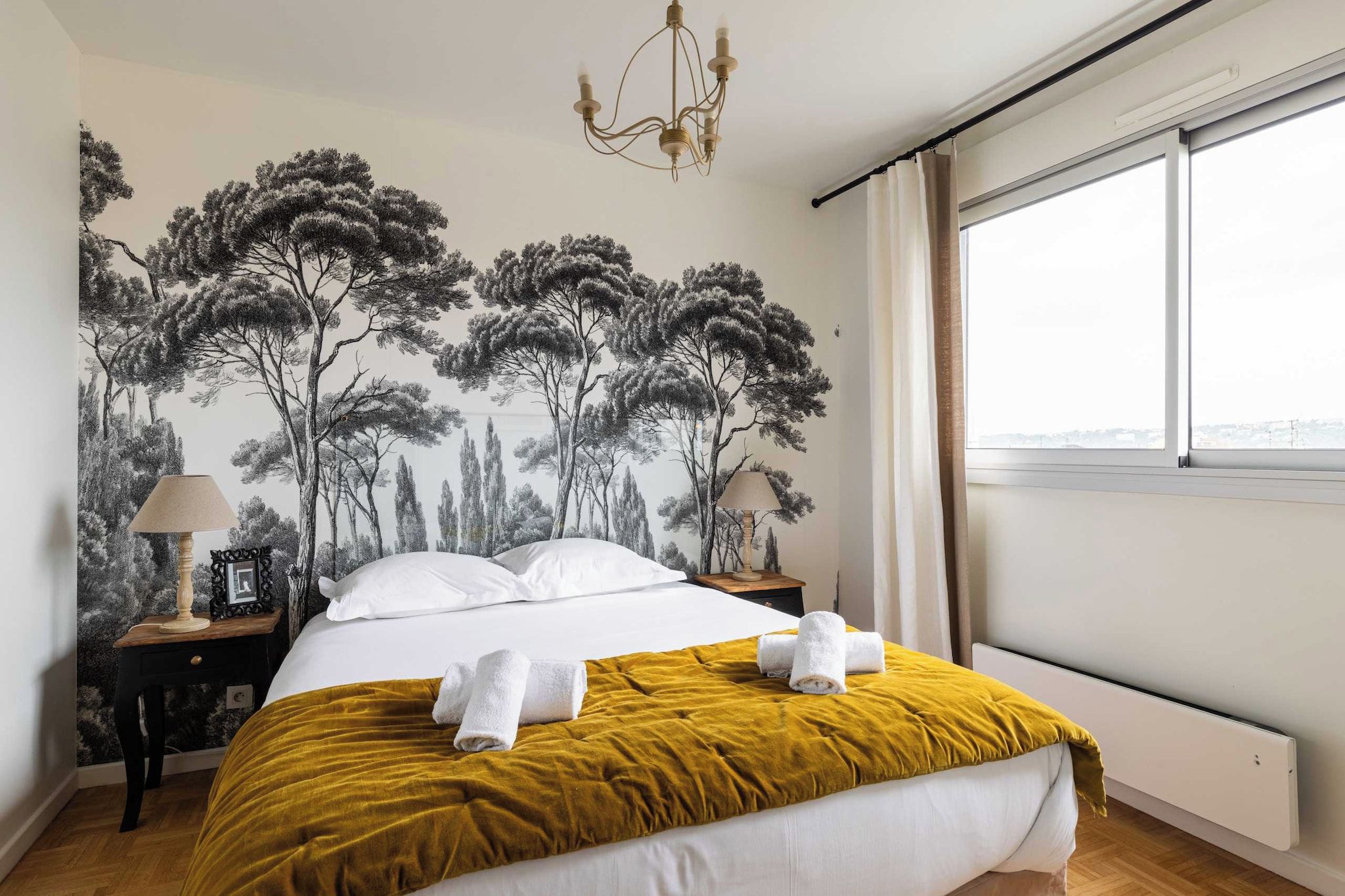 A bedroom with a bed, two nightstands, a chandelier, a window with curtains, and a wall with a tree mural.