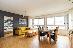 A modern living room with a yellow sofa, a dining table with chairs, and large windows.