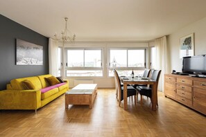 A modern living room with a yellow sofa, a wooden coffee table, a dining table with chairs, and a TV on a wooden cabinet.