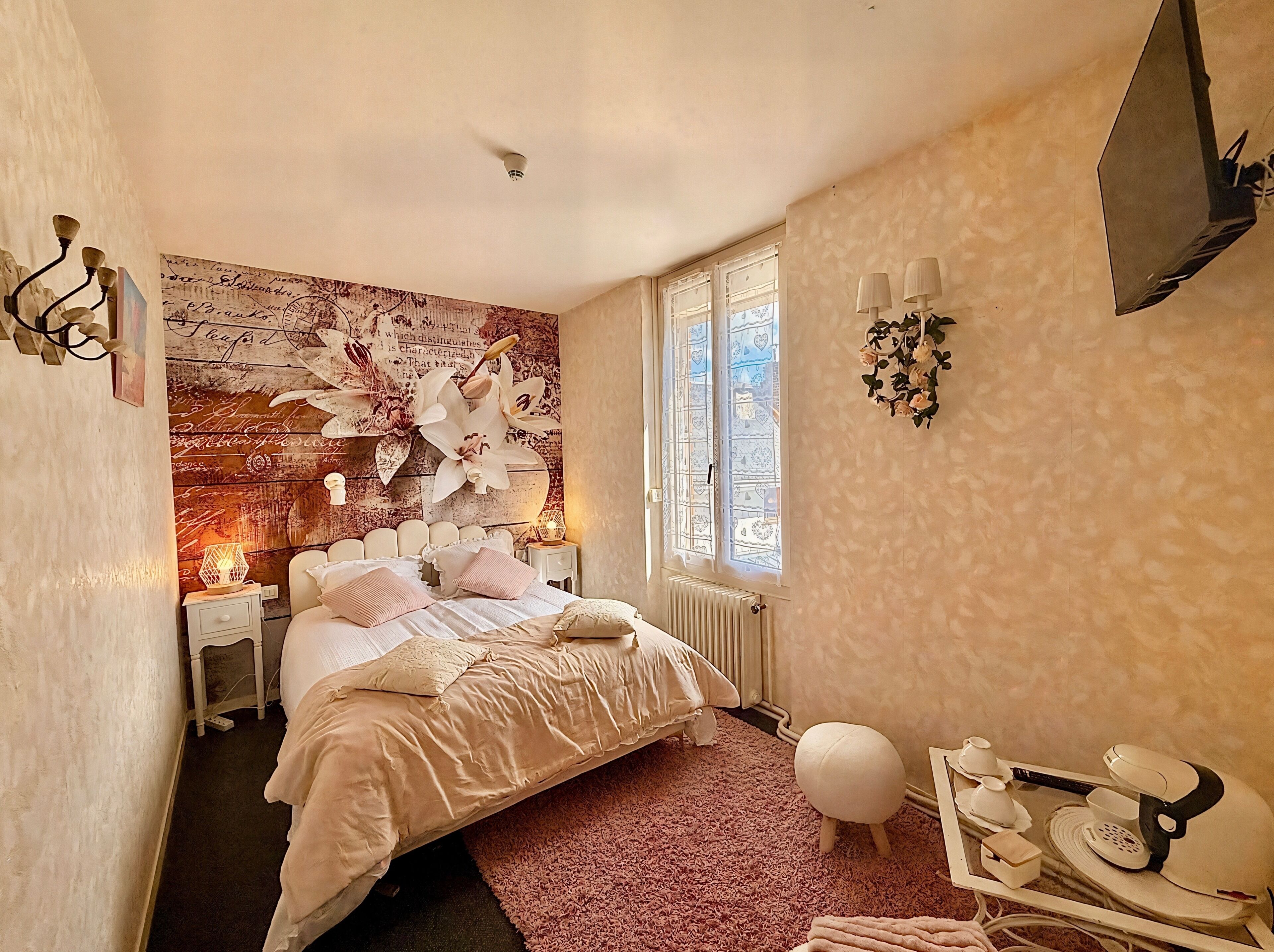 Een slaapkamer met een bed, nachtlampjes, een nachtkastje, een radiator, een raam met decoratief glas en een aan de muur bevestigde televisie.