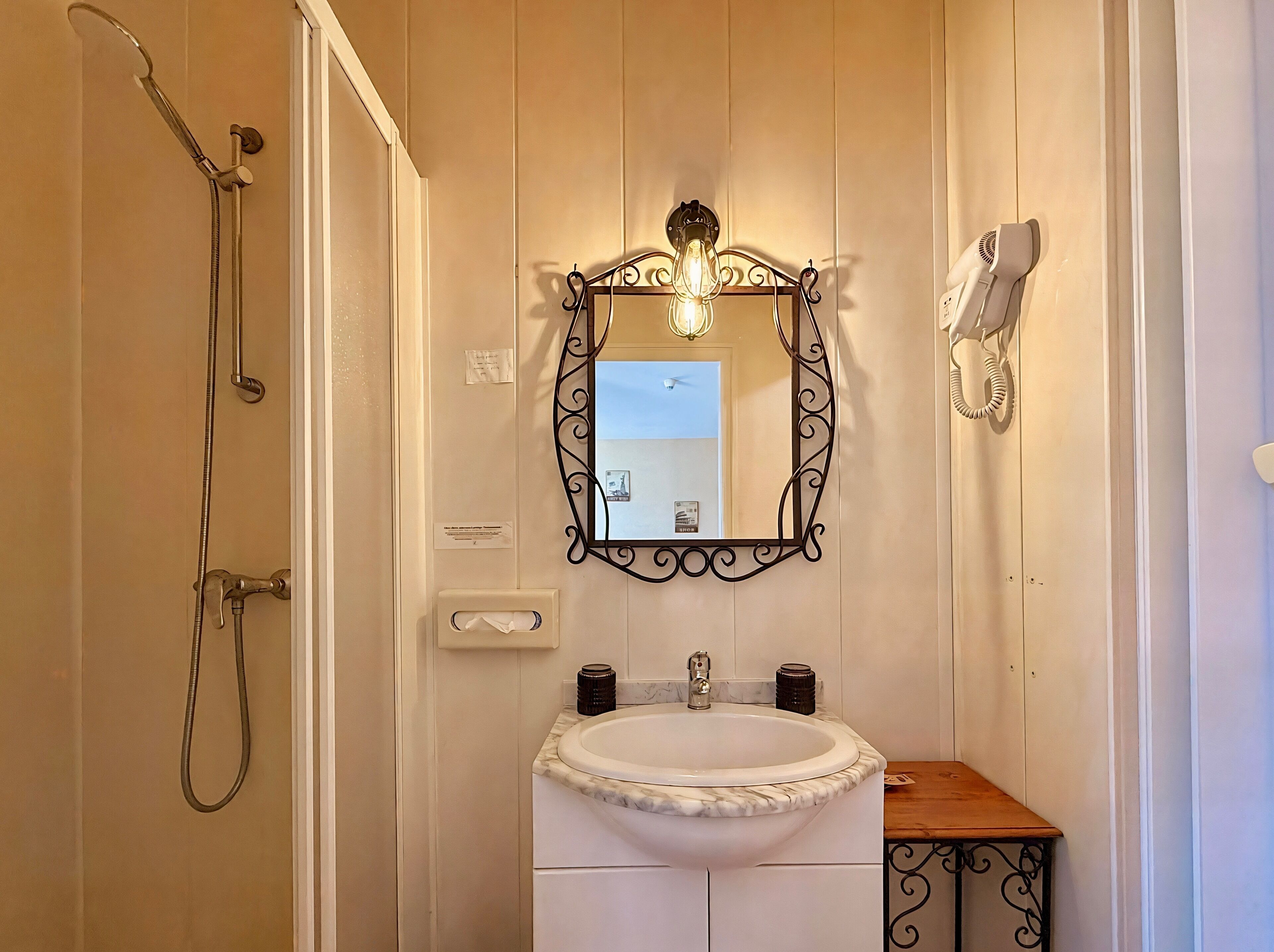 A bathroom with a shower, a sink with a marble countertop, a mirror with a decorative frame, and a hairdryer mounted on the wall.