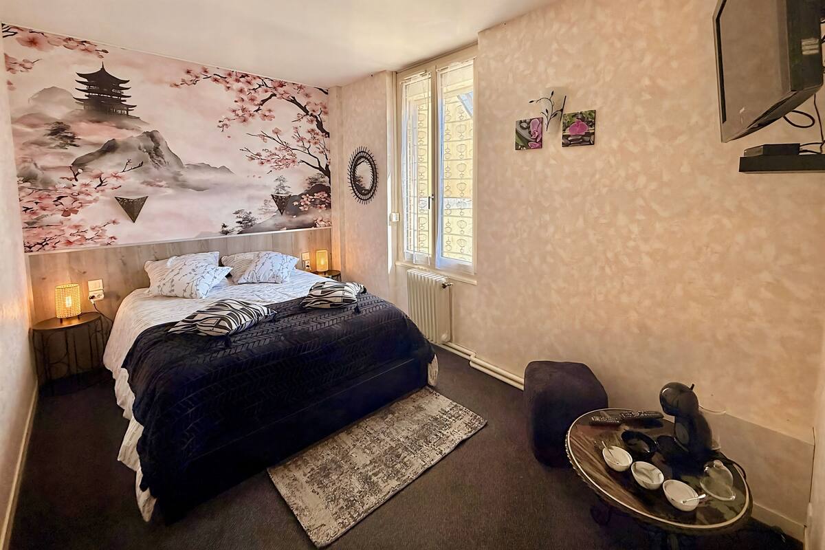 A bedroom with a bed, a nightstand with a lamp, a window with blinds, a radiator, and a small table with a remote control.