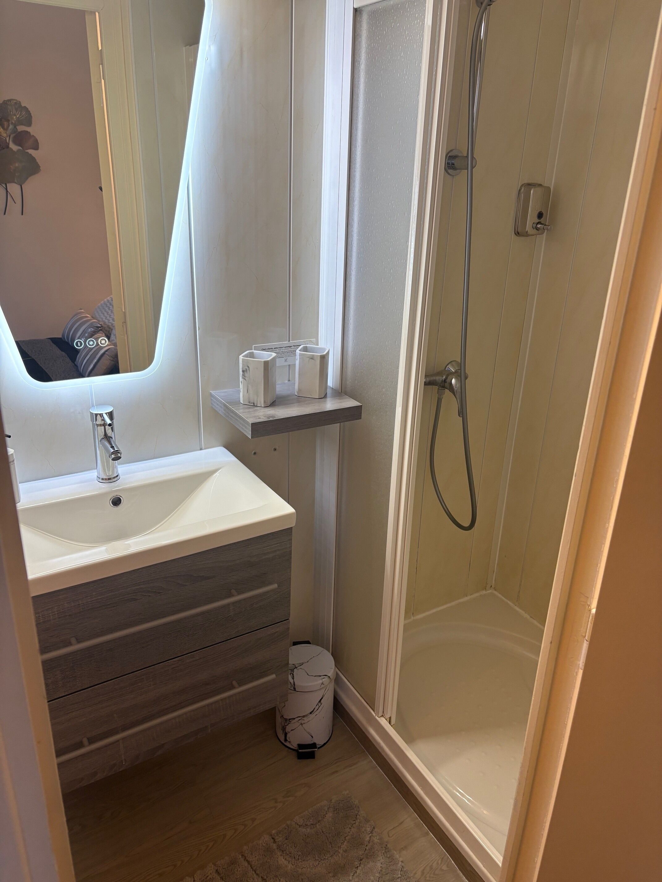 Un baño con lavamanos blanco, una lavabo de mármol, tocador de madera, ducha con puerta de cristal y un pequeño estante con dos recipientes blancos.