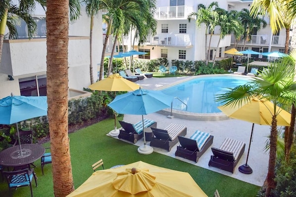 A pool area with lounge chairs and umbrellas, surrounded by palm trees and a building with balconies.