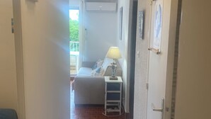 Un couloir étroit, dont le sol est recouvert de carreaux, mène à une chambre à coucher équipée d’un lit, d’une table de chevet avec une lampe et d’une fenêtre donnant sur un paysage verdoyant.