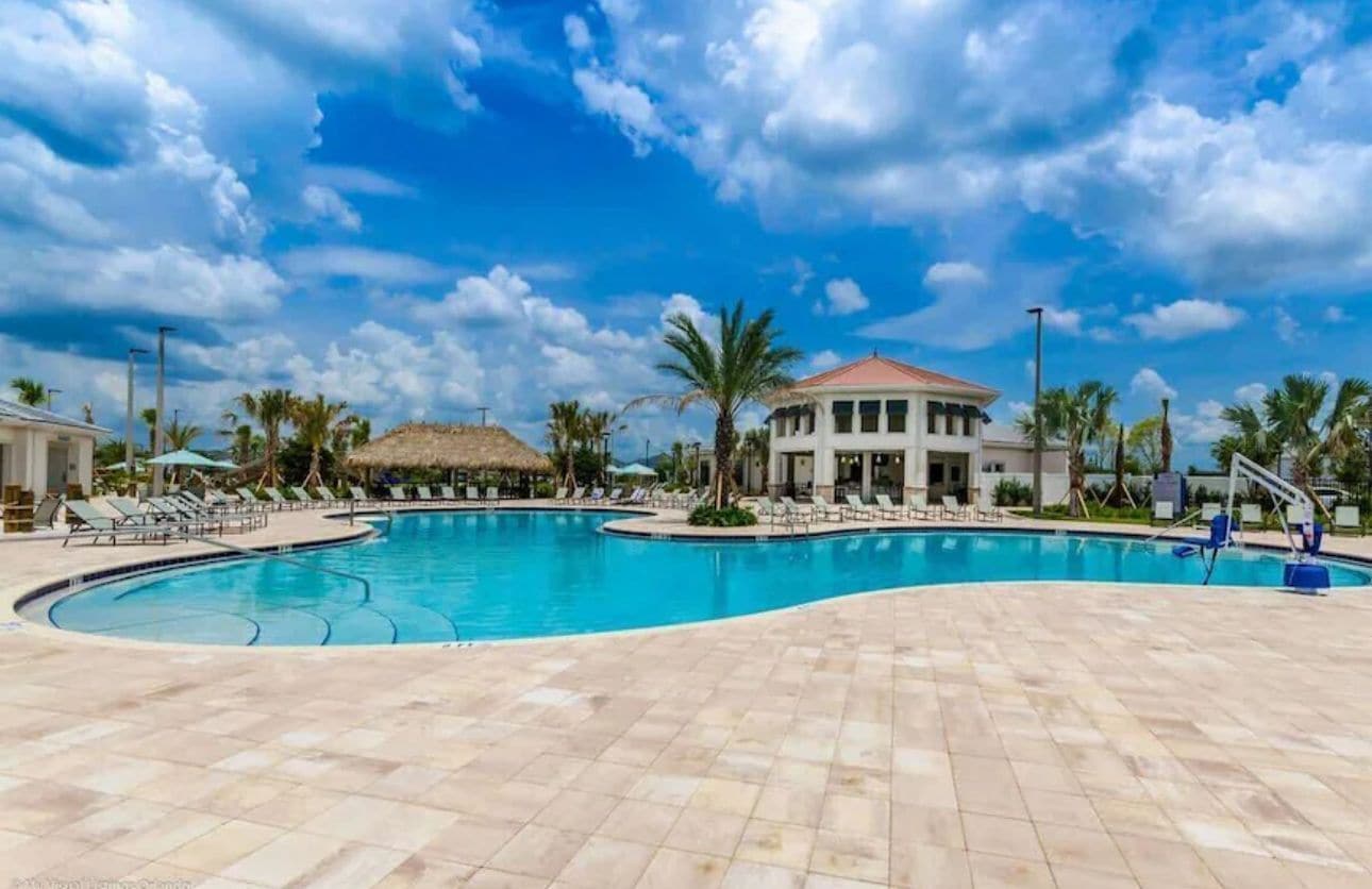 A large outdoor swimming pool with lounge chairs and palm trees, surrounded by buildings with a tropical architectural style.