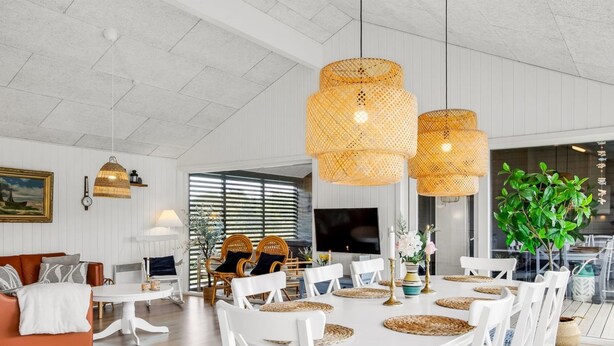Zona pranzo con tavolo bianco apparecchiato per sei, circondato da sedie bianche con cuscini blu. Tre lampade a sospensione in vimini pendono sopra il tavolo.