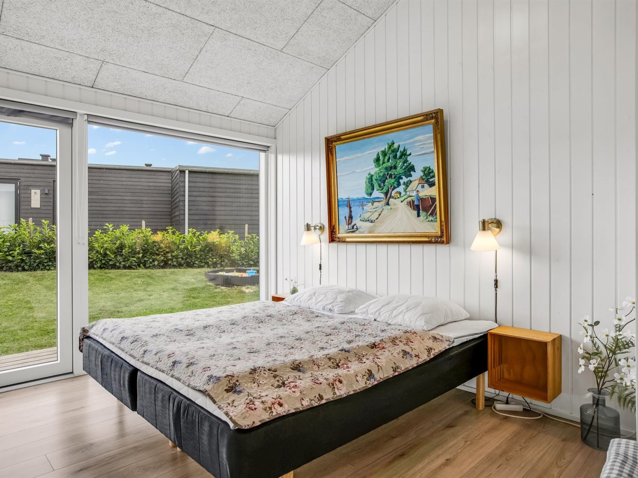 A bedroom with a bed, a painting, a nightstand, and a view of the outdoors.