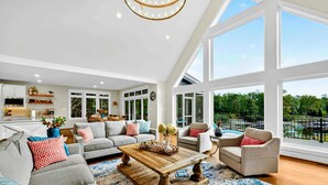 A spacious living room with a large window, a kitchen in the background, and a pool view.