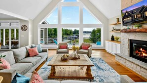 A spacious living room with a high ceiling, large windows, a fireplace, and a view of the outdoors.