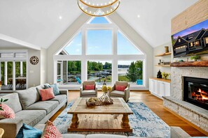 A spacious living room with a high ceiling, large windows, a fireplace, and a view of the outdoors.