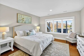 A bedroom with a large bed, two bedside tables with lamps, a dresser, a window with a view of trees, and a painting on the wall.