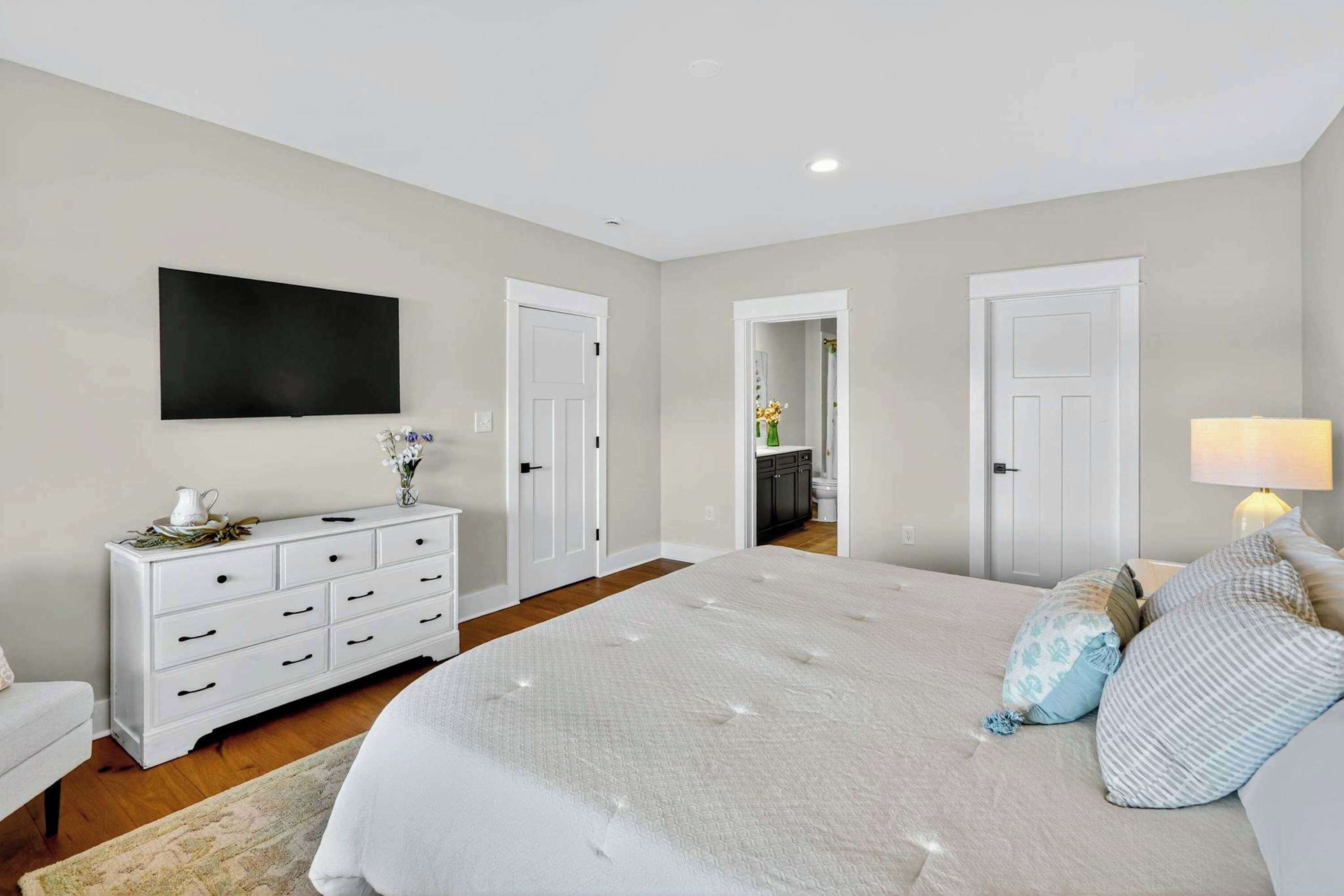 A bedroom with a large bed, a dresser, a TV, and a door leading to a bathroom.