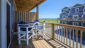 Eine Holzterrasse mit Außenmöbeln, Blick auf den Strand und ein mehrstöckiges Gebäude im Hintergrund.