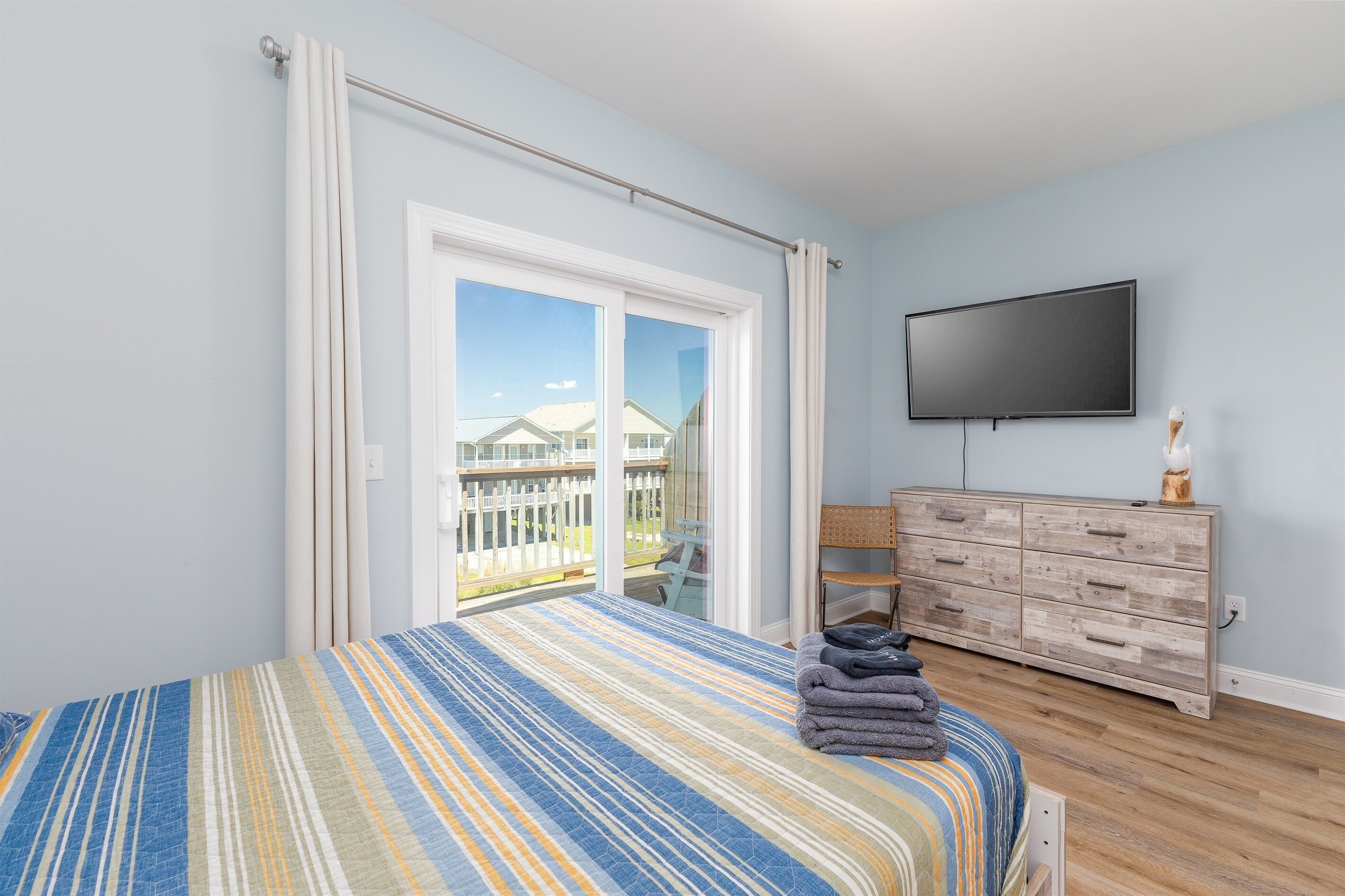 A bedroom with a large bed, a TV mounted on the wall, a wooden dresser, and a sliding glass door leading to a balcony.
