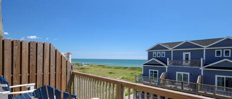 A wooden deck with blue chairs, a view of the ocean, and a two-story building with a balcony.