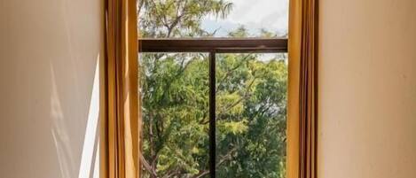 A room with a round wooden table and two chairs, a large window with curtains, and a view of greenery outside.