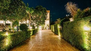 A paved pathway flanked by well-maintained hedges and trees, leading to a building with illuminated windows. - Aditya Palace (Pushkar)