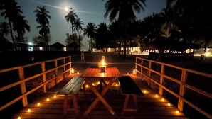 Een houten terras met banken en tafels, omgeven door palmbomen en een nachtelijke hemel.