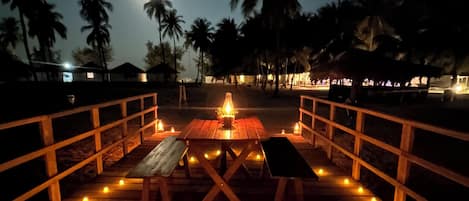 A wooden deck with benches and tables, surrounded by palm trees and a night sky.