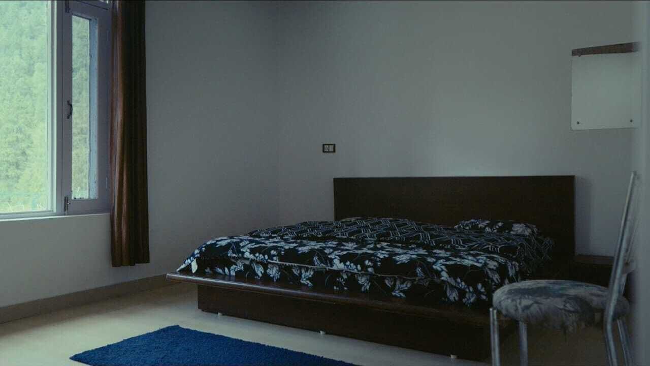 A bedroom with a bed, a chair, a window with curtains, and a wall-mounted light fixture.