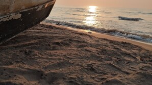 Un bote con la palabra "ANDALAS" en su lateral, posicionado en una playa arenosa con el océano y un sol poniéndose de fondo.